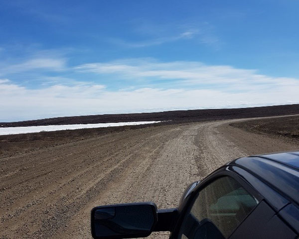 A car on the Inuvik to Tuktoyaktuk highway in the spring of 2018 after it was just opened. The highway is a gravel surface surrounded by local plants and a river. 