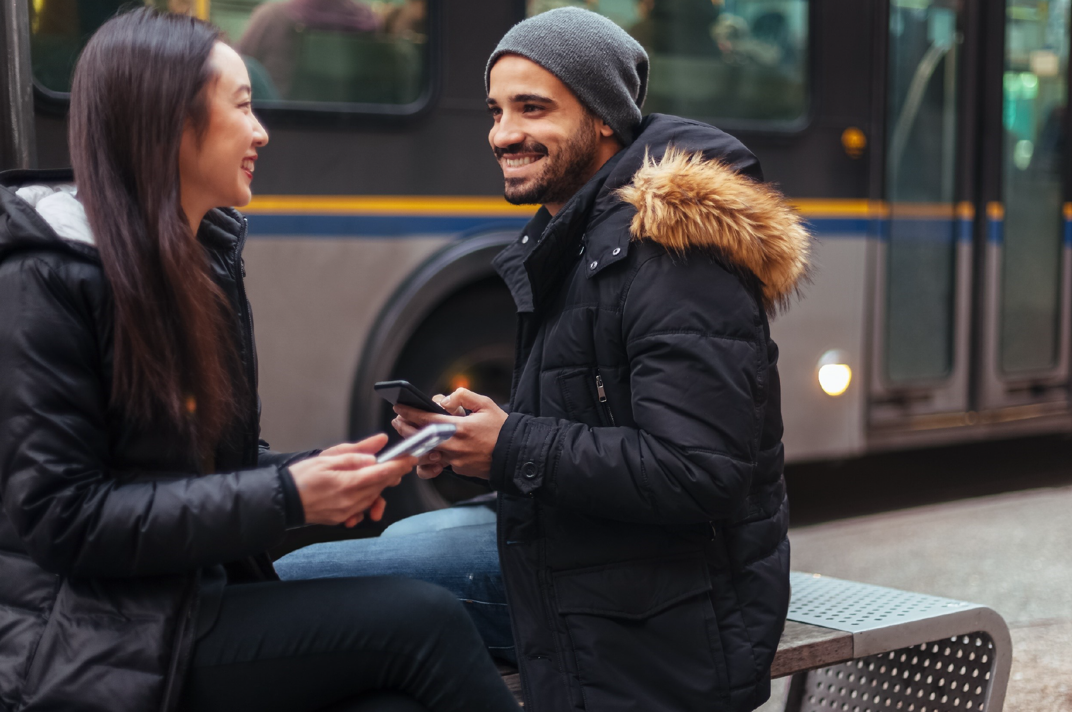 Two people smiling at each other sitting on a bench holding their phones with a bus driving by behind them.