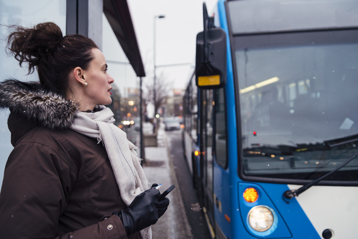 A woman looks at a bus pulling up while she holds her phone and transit pass. Snow is lightly falling.