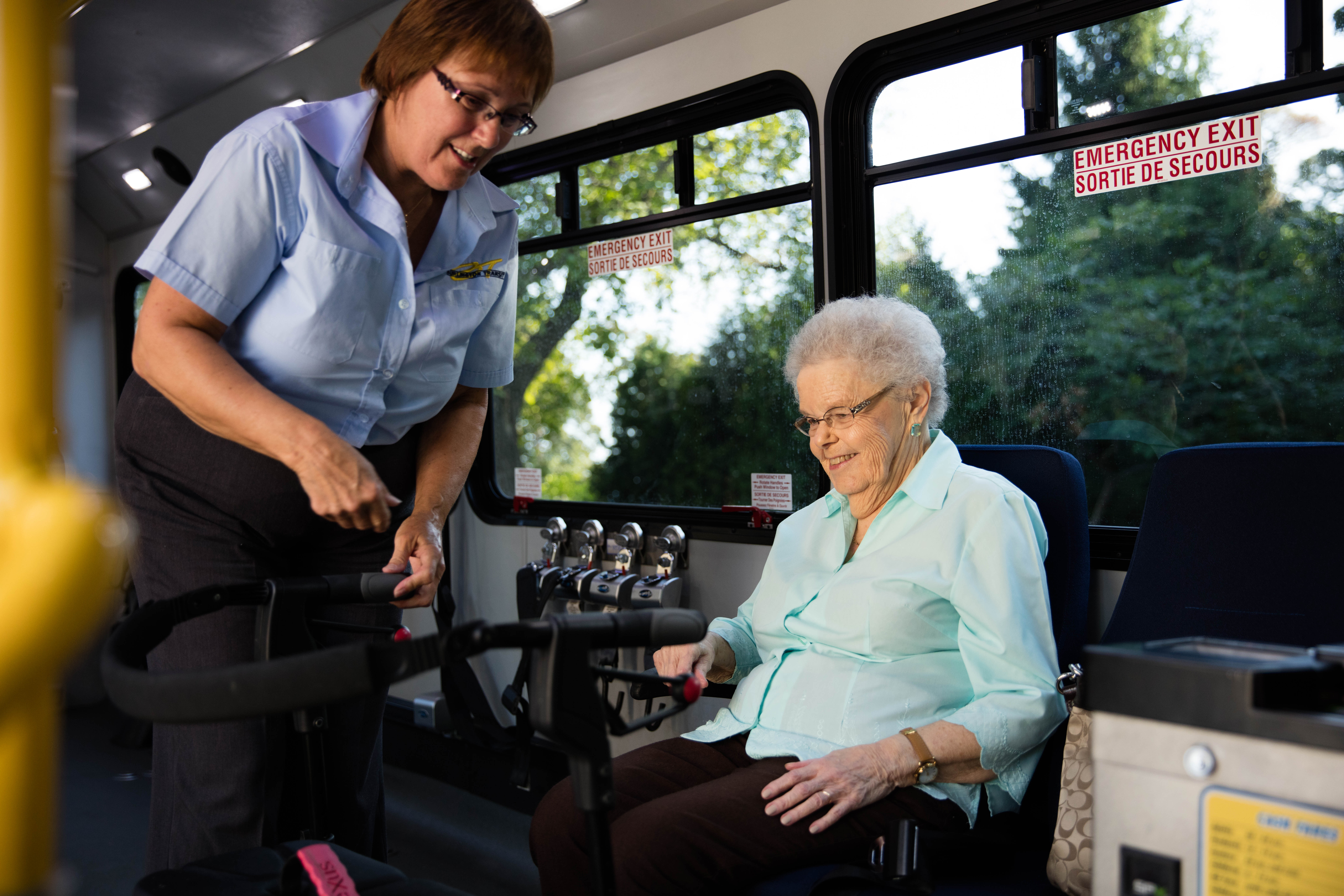 A bus operator helps a senior woman who is sitting on a bus and smiling looking down at her walker.